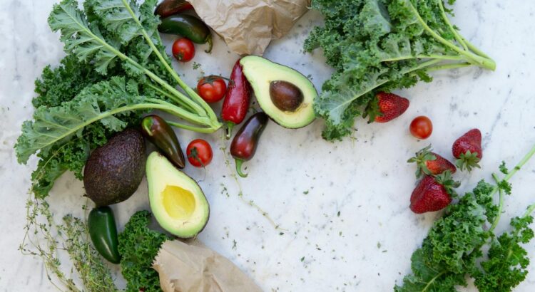 Top view of fresh avocados, kale, tomatoes, and peppers arranged on a marble surface.